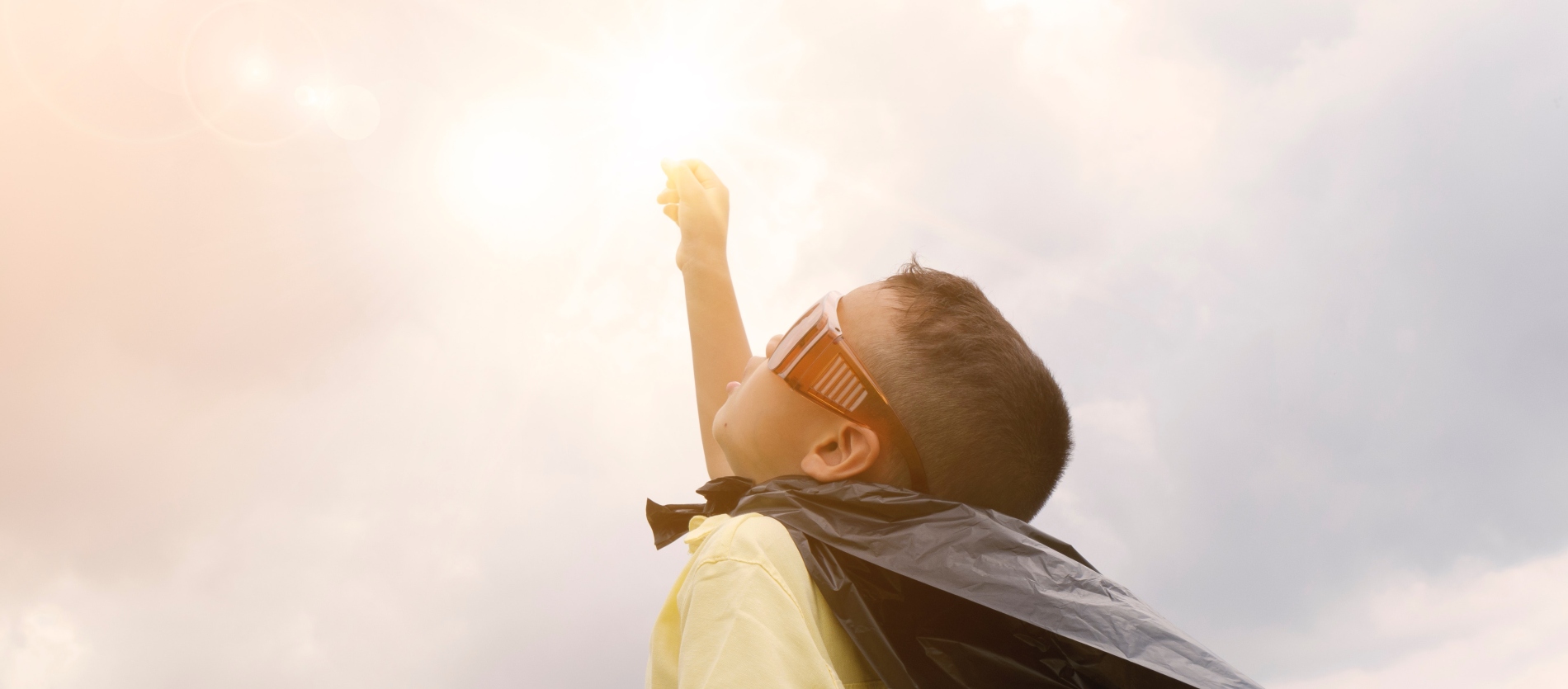 Young boy wearing sunglasses and a black cape stretching his right arm up towards the sun